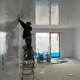 TM Milena Tomczak - Dwóch pracowników na drabinie aplikuje maszynowo gładź na ścianę w nowym mieszkaniu, widoczny agregat malarski i wiadro z farbą.