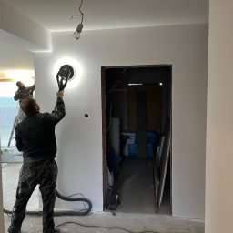 TM Milena Tomczak - Prace wykończeniowe w pomieszczeniu: szlifowanie ściany przy użyciu szlifierki z odkurzaczem, widoczny pracownik w kombinezonie, surowy stan w sąsiednim pomieszczeniu, wisząca żarówka na kablu.