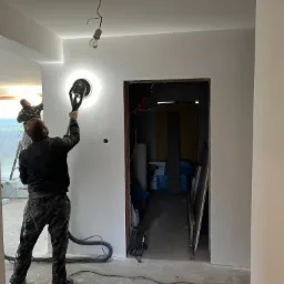 Prace wykończeniowe w pomieszczeniu: szlifowanie ściany przy użyciu szlifierki z odkurzaczem, widoczny pracownik w kombinezonie, surowy stan w sąsiednim pomieszczeniu, wisząca żar&oacute;wka na kablu.