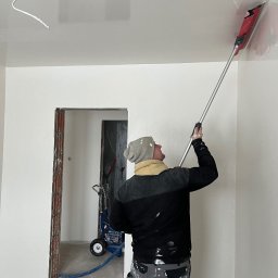 TM Milena Tomczak - Pracownik w czapce i roboczych spodniach, z tyłu, szpachluje sufit za pomocą narzędzia z długim uchwytem; w tle widoczny agregat malarski i surowa ościeżnica drzwi.