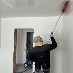 Pracownik w czapce i roboczych spodniach, z tyłu, szpachluje sufit za pomocą narzędzia z długim uchwytem; w tle widoczny agregat malarski i surowa ościeżnica drzwi.