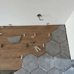 Układanie szarych, heksagonalnych płytek na podłodze, częściowo na drewnopodobnym podkładzie, z użyciem białych krzyżyków dystansowych, widoczne niedociągnięcia przy ścianie i wystające kable...
