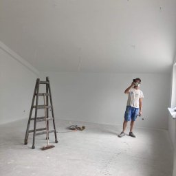 TM Milena Tomczak - Wnętrze świeżo pomalowanego pokoju z drabiną, narzędziami i mężczyzną w masce ochronnej robiącym zdjęcie telefonem; widoczne ślady na podłodze.