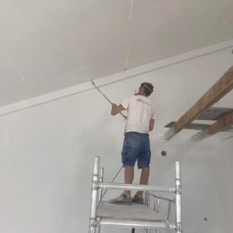 TM Milena Tomczak - Pracownik maluje sufit natryskowo z rusztowania, widoczne kable wystające z sufitu i drewniane elementy konstrukcyjne.
