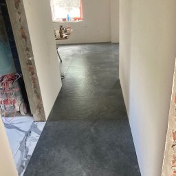 TM Milena Tomczak - Korytarz w trakcie prac wykończeniowych: szare płytki podłogowe ułożone wzdłuż, ściany przygotowane do malowania, widoczne surowe fragmenty muru i narzędzia w tle.