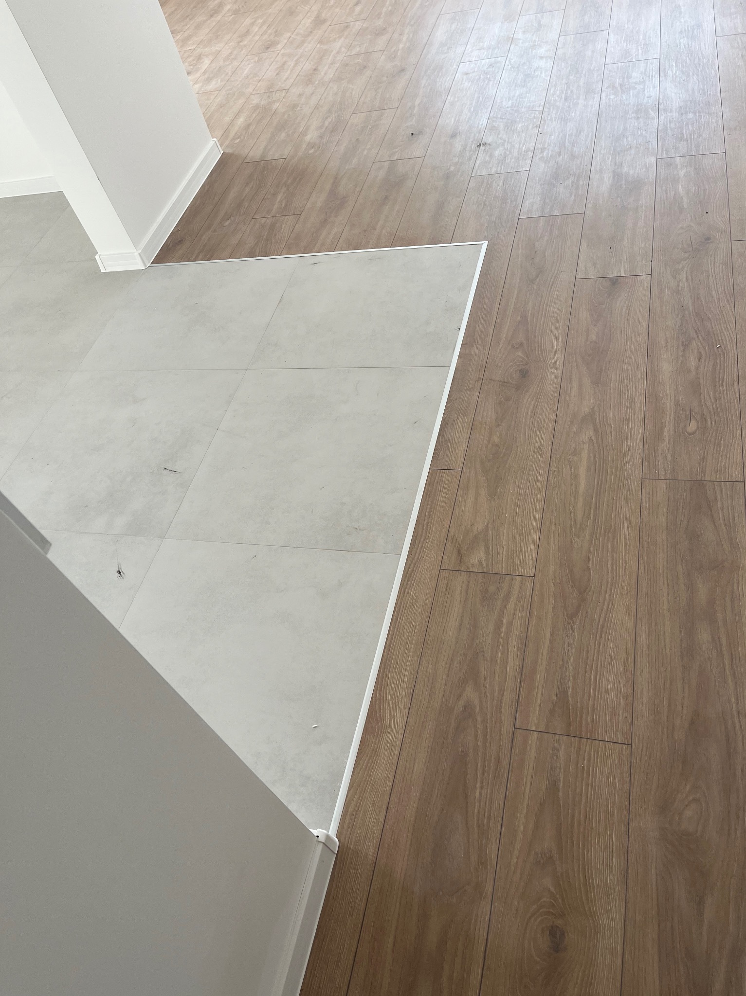 Połączenie jasnej, kwadratowej posadzki z drewnopodobnymi panelami, widoczne listwy przypodłogowe przy ścianie.