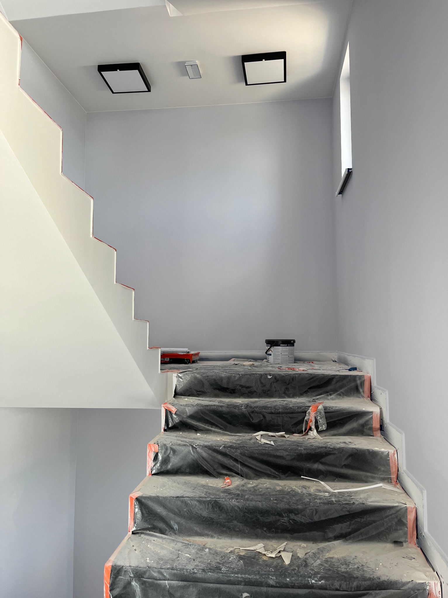 Schody zabezpieczone folią malarską podczas prac wykończeniowych, widoczna puszka farby i kuweta malarska, świeżo pomalowane ściany w odcieniach szarości, nowoczesne oświetlenie sufitowe...