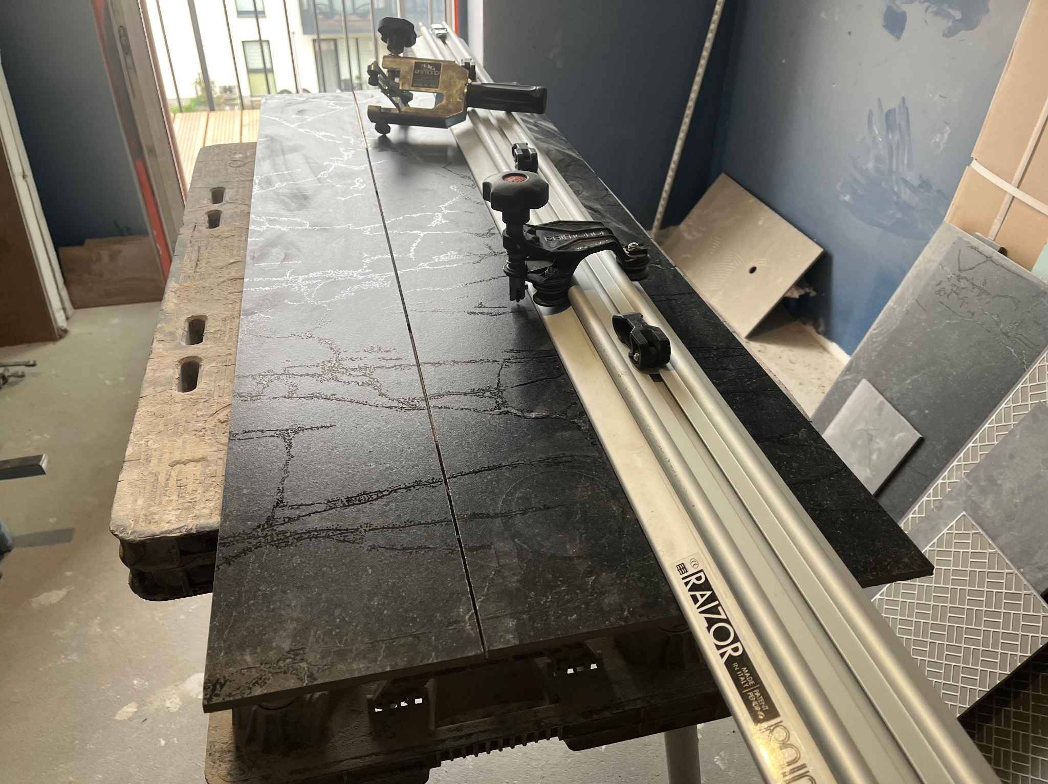Ciemnoszara płytka z marmurkowym wzorem, przecięta profesjonalną przecinarką do płytek Raimondi Razor na warsztatowym stole, w tle fragment ściany i inne płytki.