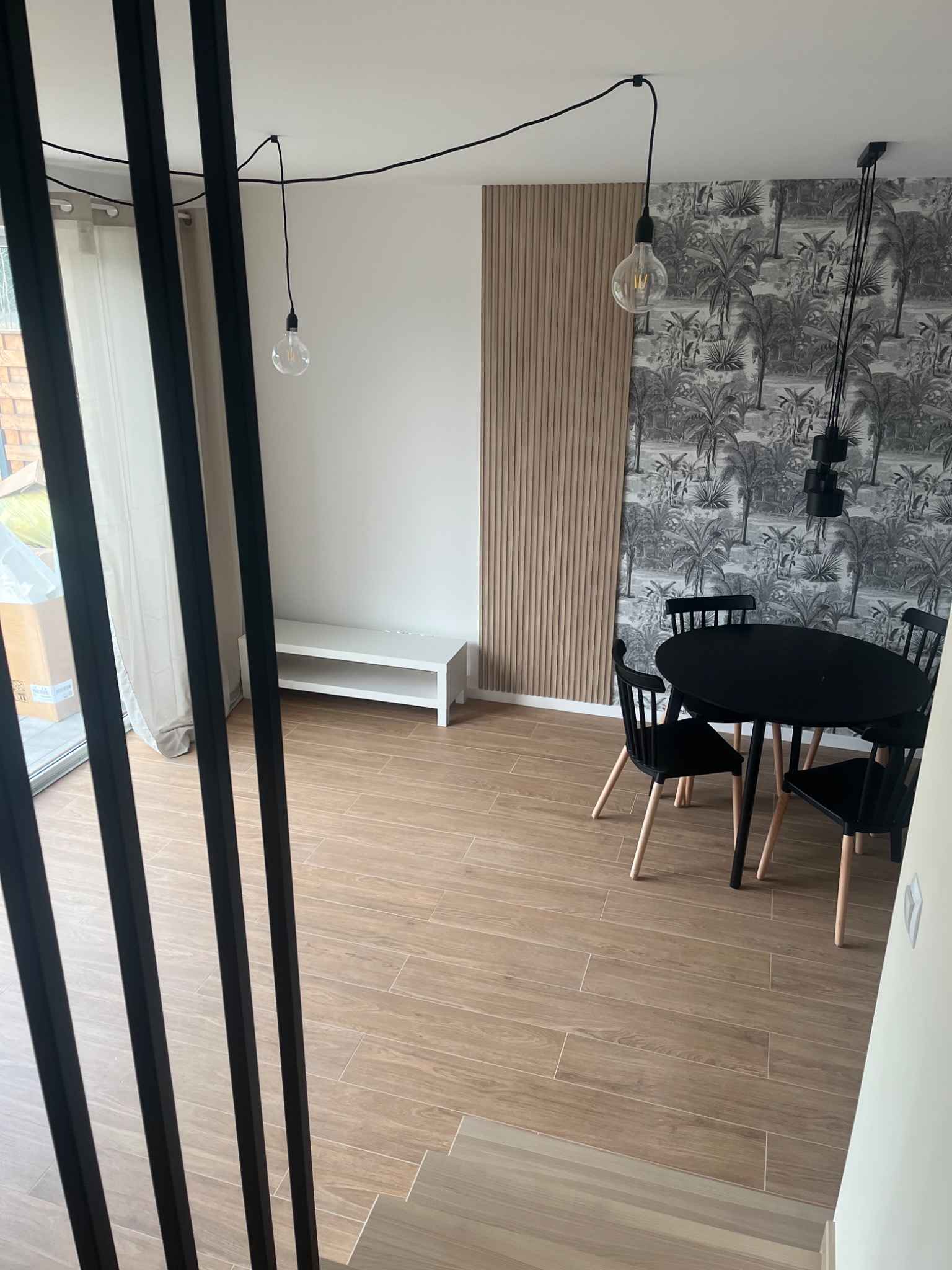 Jasne wnętrze jadalni z drewnopodobną podłogą, czarnym okrągłym stołem i krzesłami, drewnianą ścianą z pionowych paneli oraz tapetą w tropikalne motywy, oświetlone wiszącymi lampami z żarówkami...