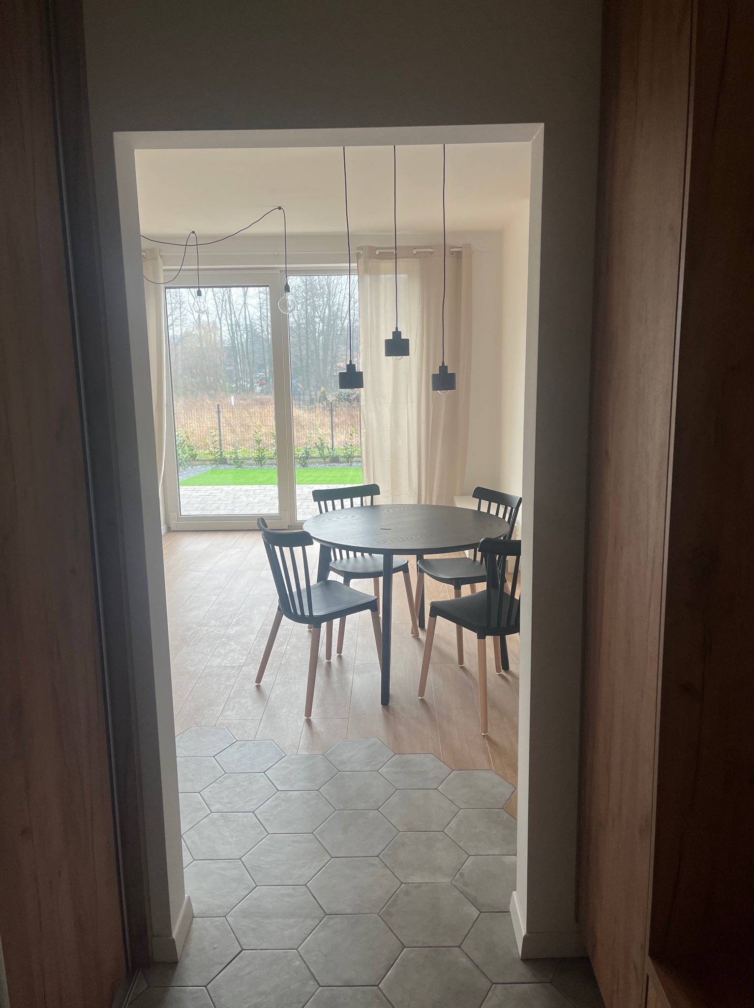 Wnętrze jadalni z okrągłym stołem i krzesłami, widok z korytarza, heksagonalne płytki przechodzące w drewnianą podłogę, minimalistyczne lampy wiszące.