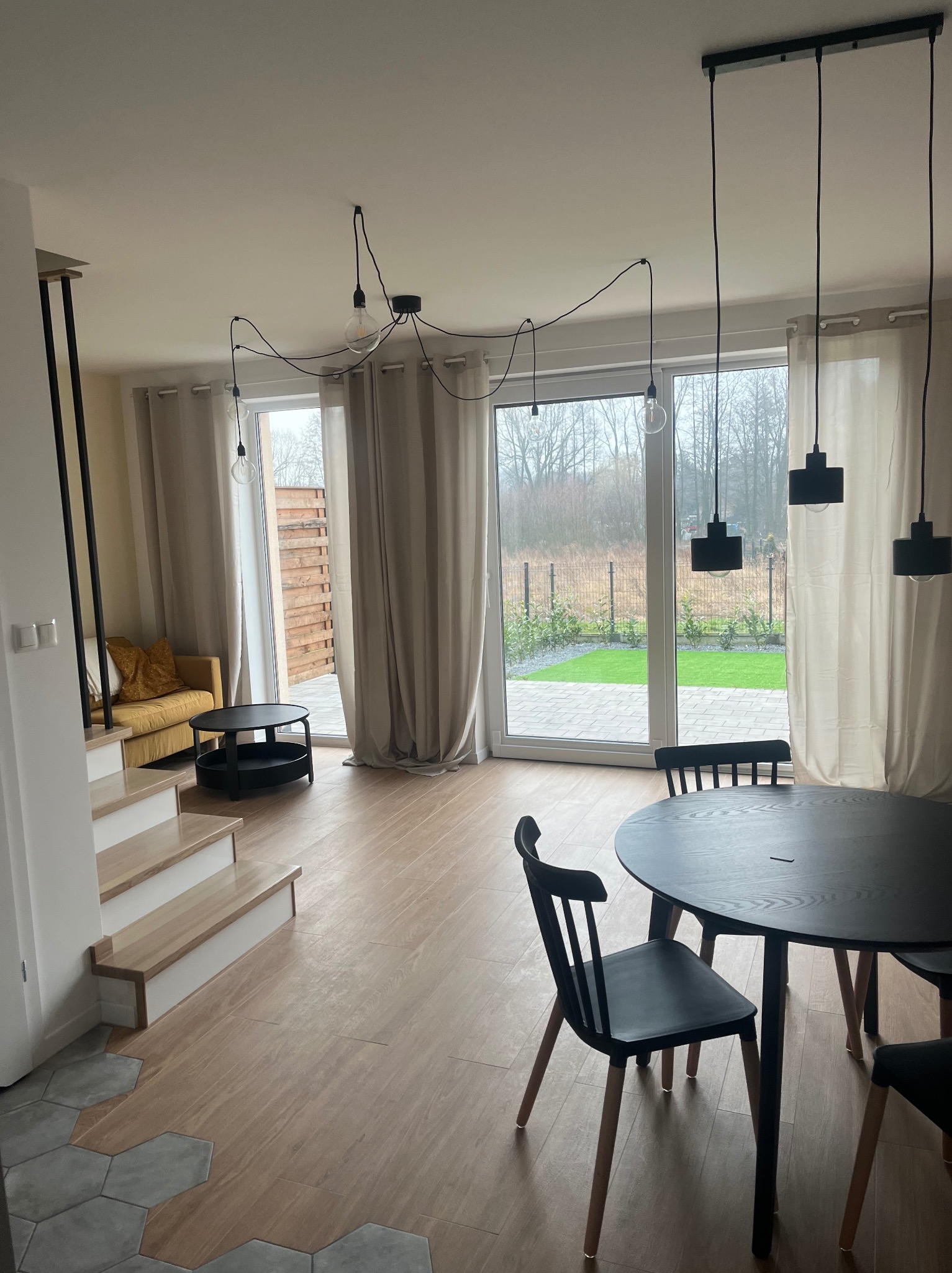 Jasne wnętrze salonu z drewnianą podłogą, schodami z drewnianymi stopniami, żółtą sofą, okrągłym czarnym stołem z krzesłami i nowoczesnymi lampami wiszącymi z żarówkami na kablach oraz trzema...