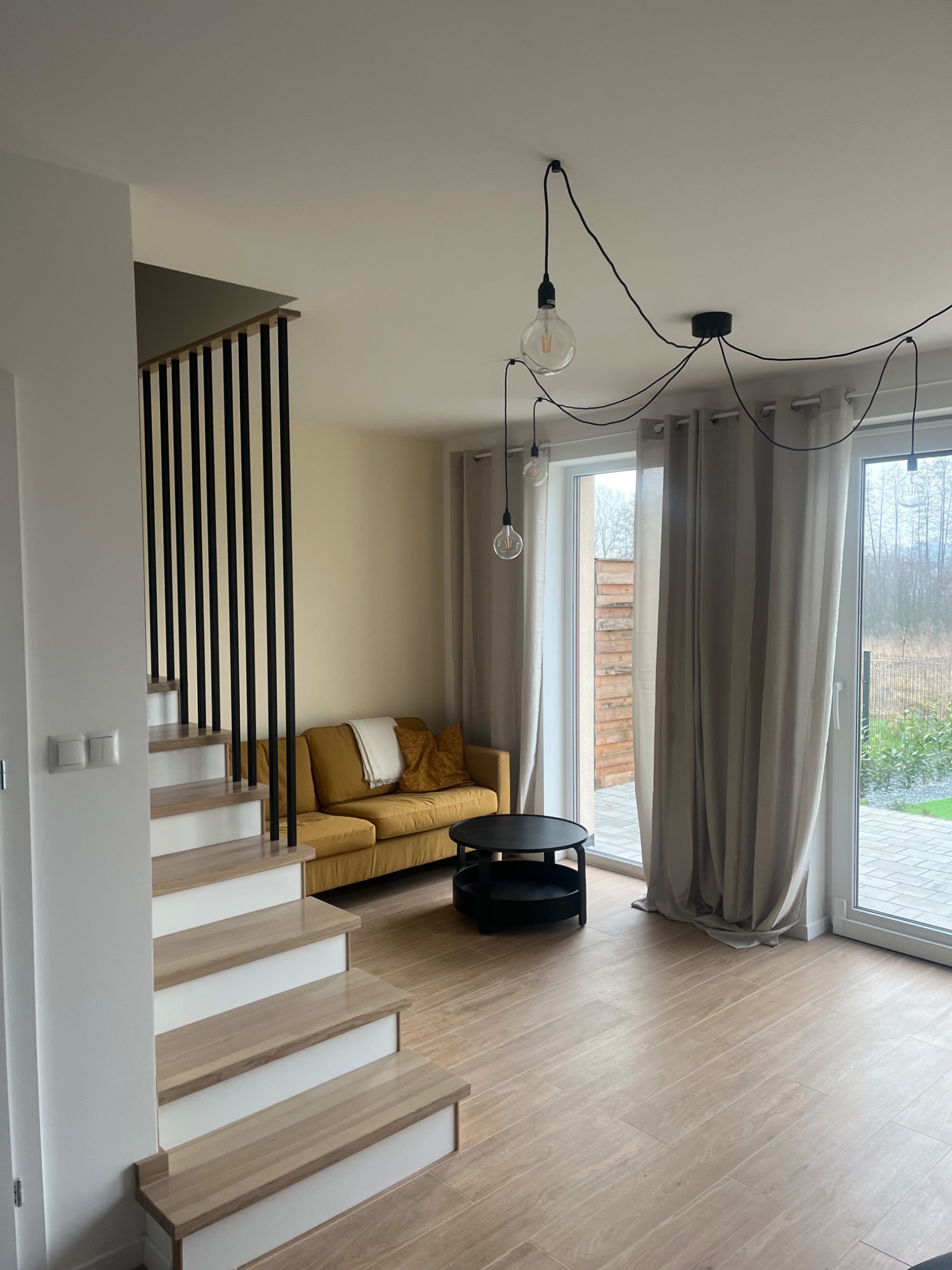 Jasne wnętrze domu z drewnianą podłogą, schodami z białymi podstopnicami, żółtą sofą, czarnym stolikiem kawowym i minimalistycznym oświetleniem zwisającym z sufitu na tle okna tarasowego.