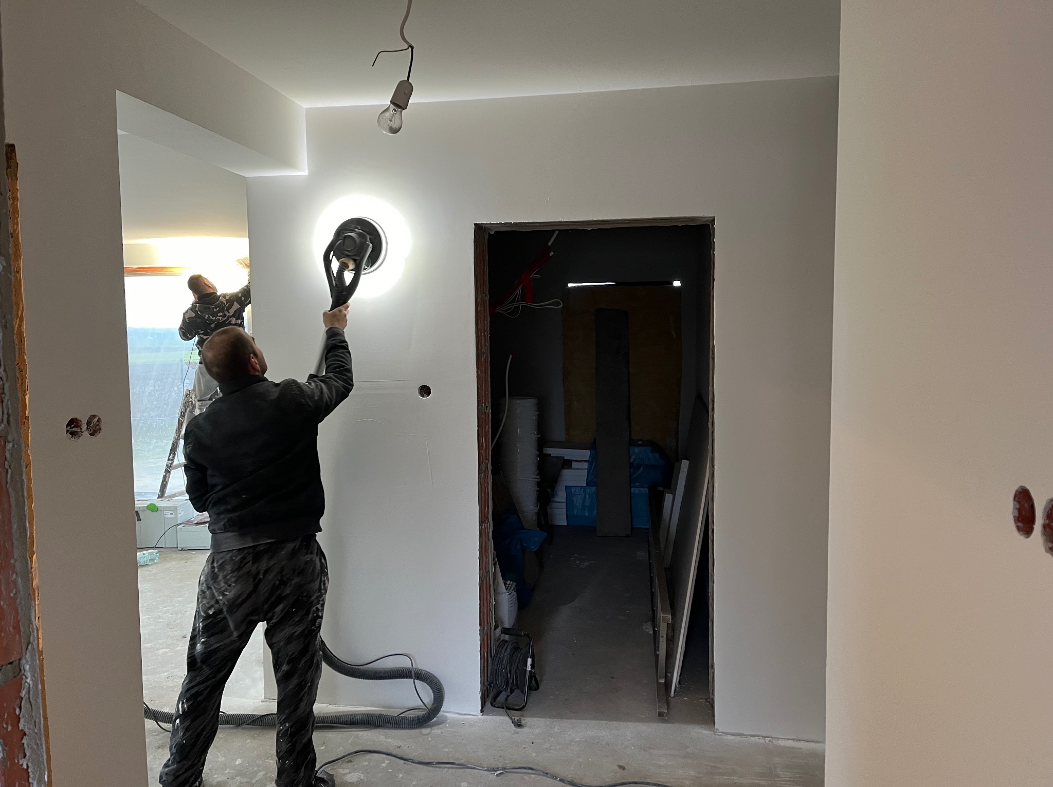 Prace wykończeniowe w pomieszczeniu: szlifowanie ściany przy użyciu szlifierki z odkurzaczem, widoczny pracownik w kombinezonie, surowy stan w sąsiednim pomieszczeniu, wisząca żarówka na kablu.