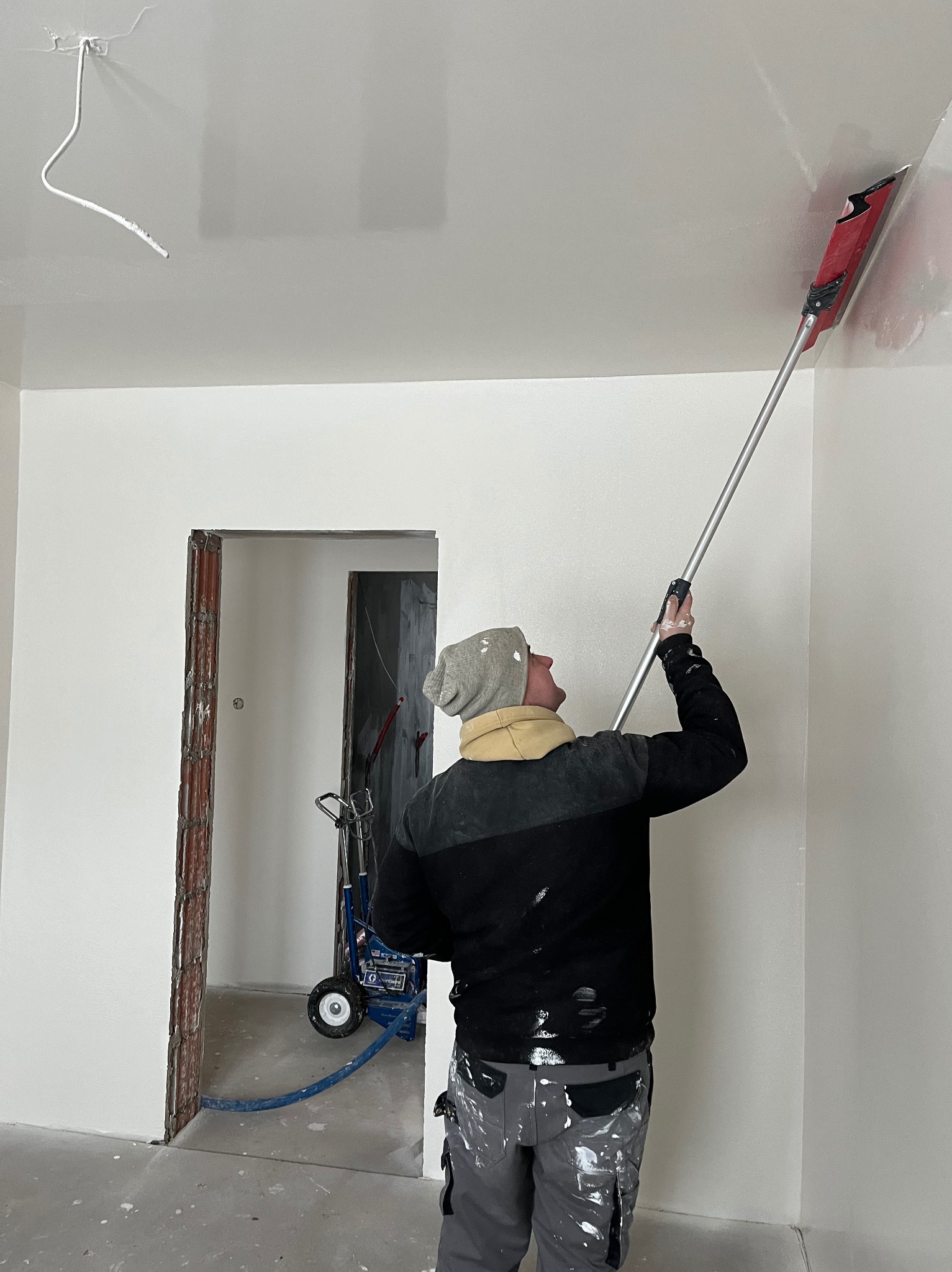 Pracownik w czapce i roboczych spodniach, z tyłu, szpachluje sufit za pomocą narzędzia z długim uchwytem; w tle widoczny agregat malarski i surowa ościeżnica drzwi.