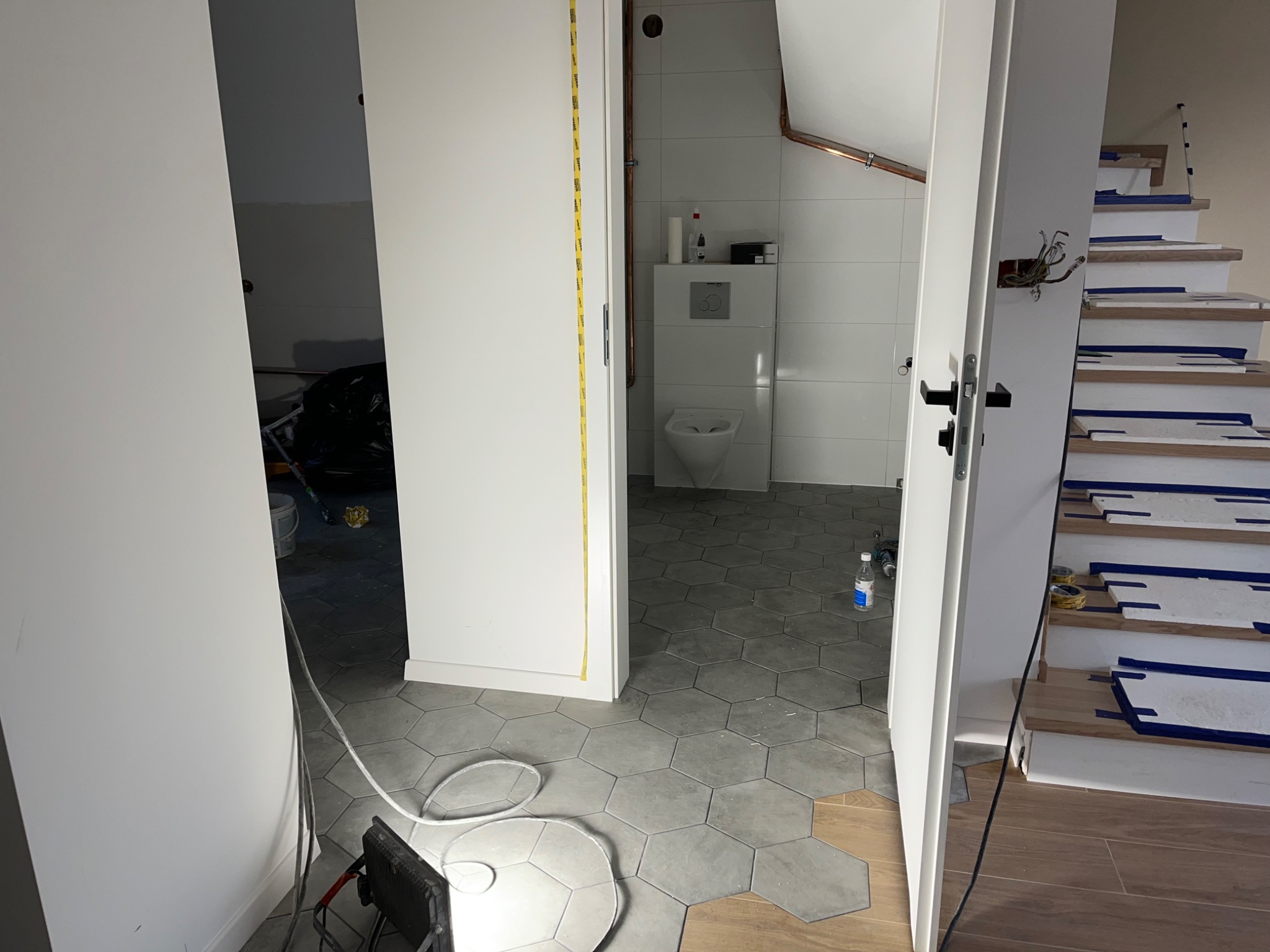Wnętrze w trakcie prac: heksagonalna szara podłoga, częściowo ułożona, widok na białą łazienkę z toaletą i schody zabezpieczone taśmą malarską.