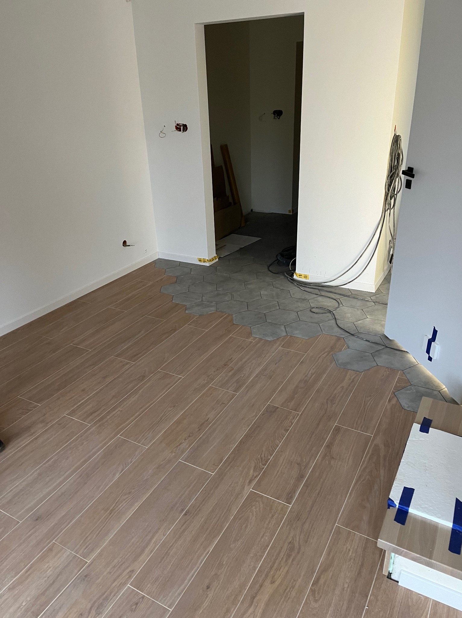 Wnętrze w trakcie prac: układanie podłogi z płytek imitujących drewno przechodzących w szare heksagonalne płytki, widoczne przygotowane płytki, kable elektryczne i surowe ściany.