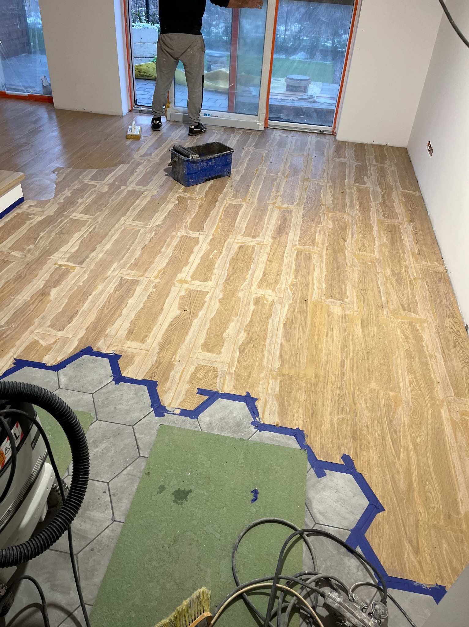 W trakcie układania drewnianego parkietu w jodełkę, widoczne resztki kleju i taśma malarska zabezpieczająca połączenie z szarymi płytkami heksagonalnymi; w tle osoba w szarych spodniach i czarnej...