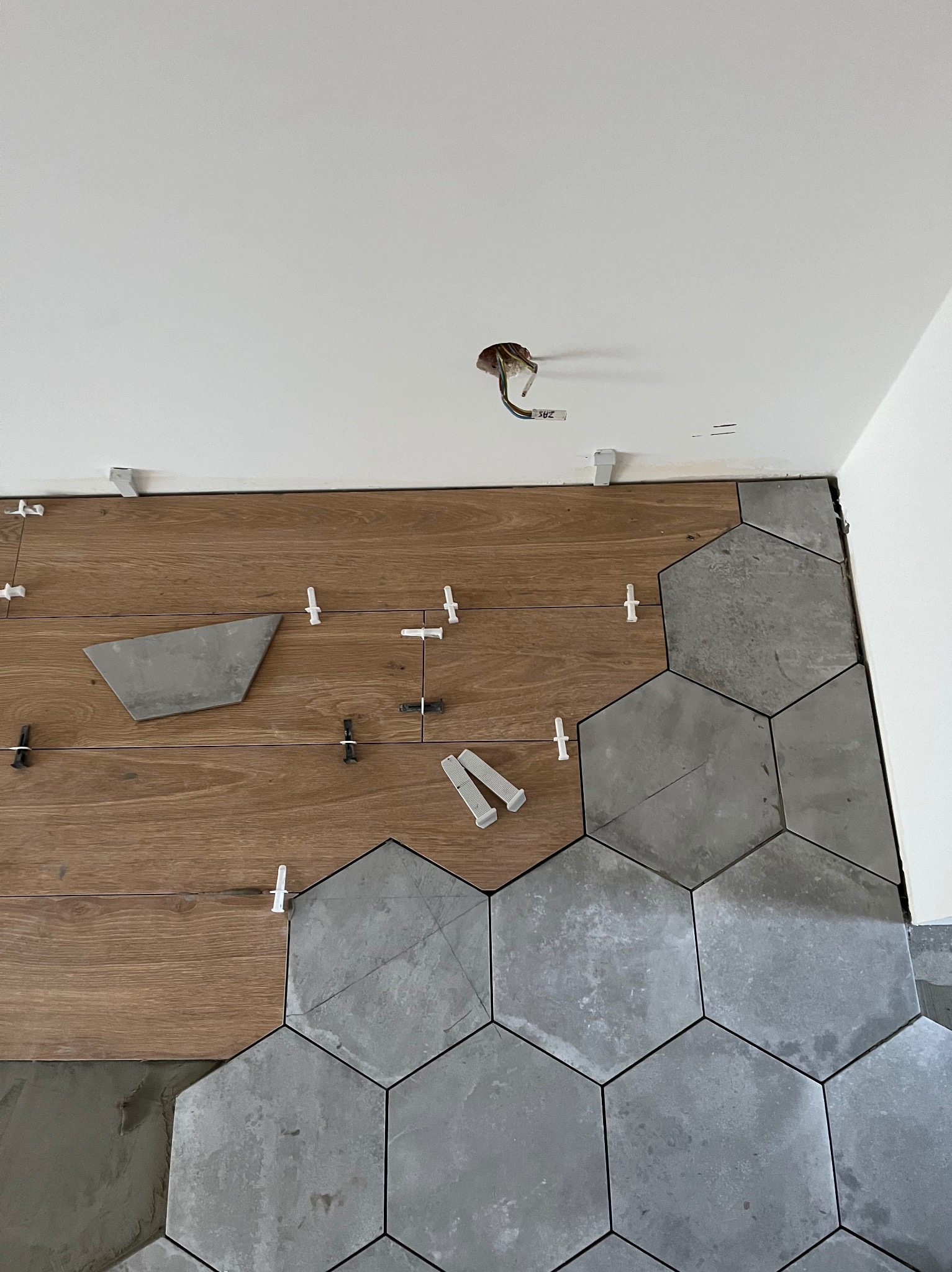 Układanie szarych, heksagonalnych płytek na podłodze, częściowo na drewnopodobnym podkładzie, z użyciem białych krzyżyków dystansowych, widoczne niedociągnięcia przy ścianie i wystające kable...
