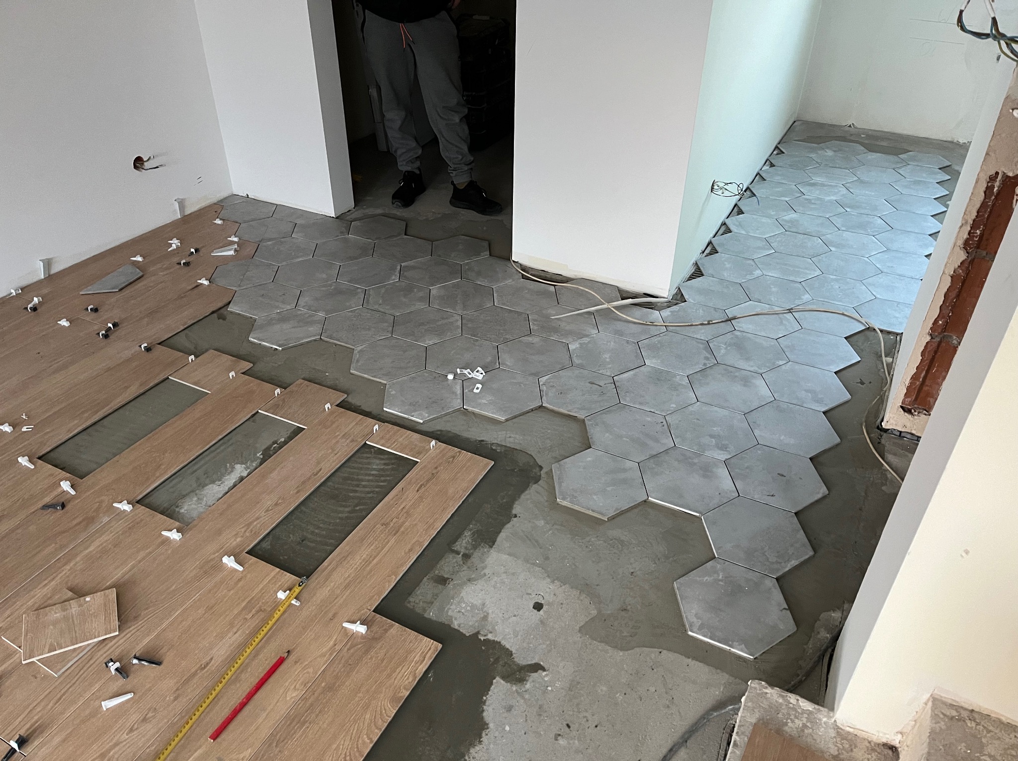 Układanie heksagonalnych szarych płytek oraz drewnopodobnych paneli podłogowych w trakcie prac wykończeniowych, widoczne krzyżyki dystansowe, poziomica i ołówek.