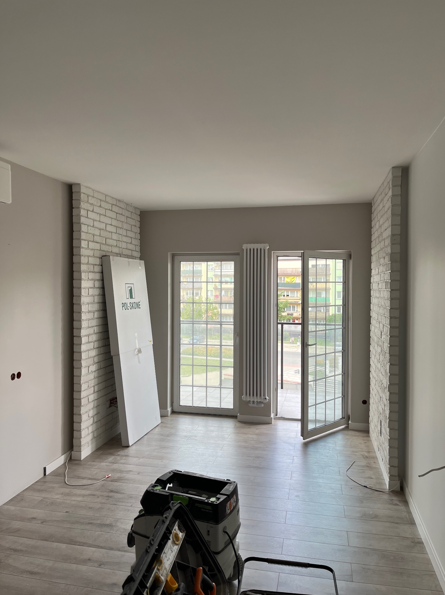 Jasne, przestronne wnętrze w trakcie prac wykończeniowych: widoczne fragmenty cegieł na ścianach, białe, przeszklone drzwi balkonowe, podłoga imitująca drewno, odkurzacz przemysłowy Festool i drzwi...