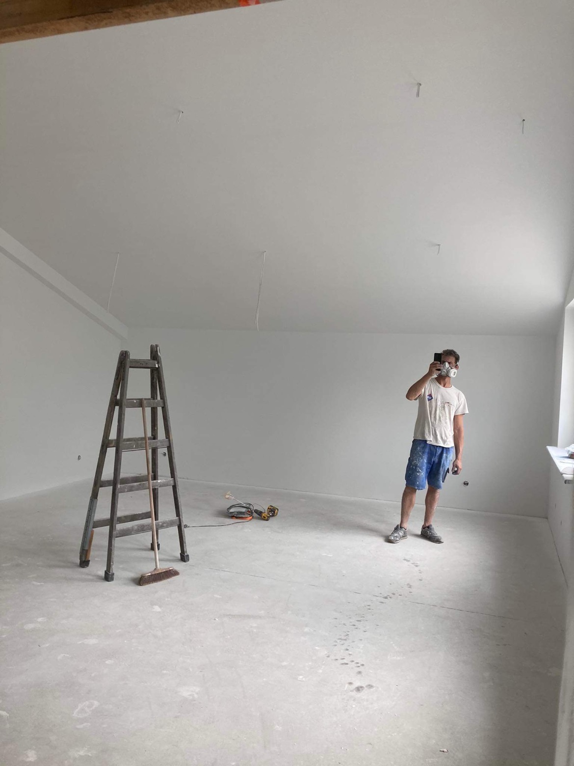 Wnętrze świeżo pomalowanego pokoju z drabiną, narzędziami i mężczyzną w masce ochronnej robiącym zdjęcie telefonem; widoczne ślady na podłodze.