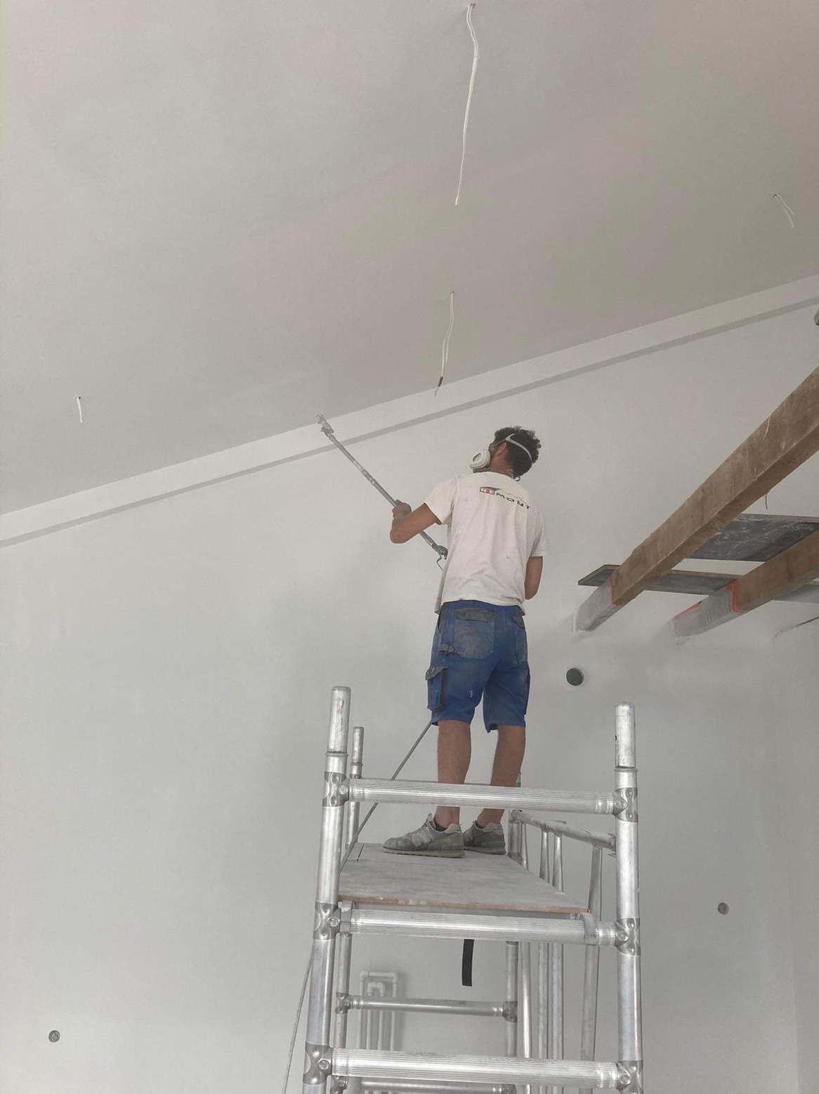 Pracownik maluje sufit natryskowo z rusztowania, widoczne kable wystające z sufitu i drewniane elementy konstrukcyjne.
