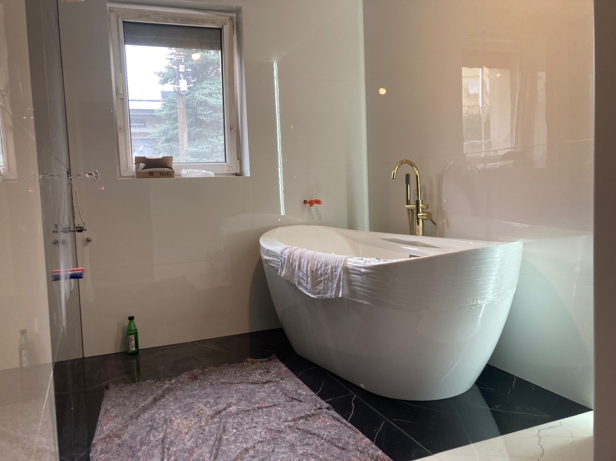 Nowoczesna biała wanna wolnostojąca z złotą baterią, zabezpieczona folią ochronną, na tle błyszczących białych płytek i czarnej podłogi z marmurowym wzorem. Okno z widokiem na zewnątrz. Na podłodze...