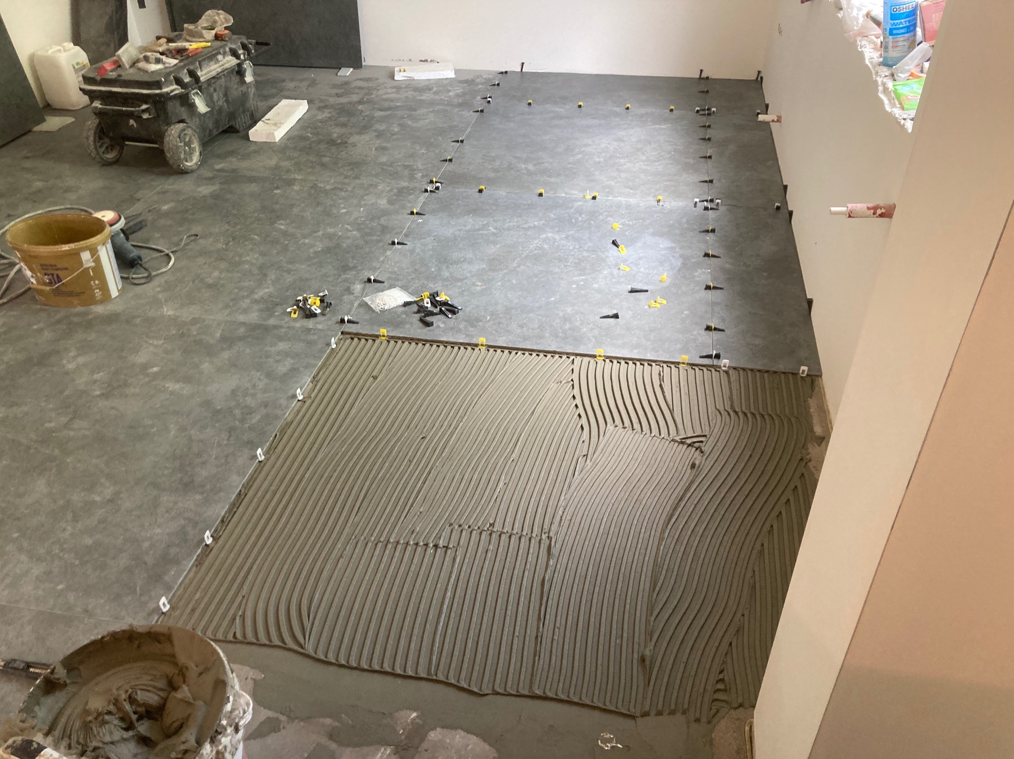 Podłoga w trakcie układania płytek, widoczna warstwa kleju z charakterystycznymi rowkami, płytki ułożone z użyciem systemu poziomowania, narzędzia i akcesoria budowlane w tle.