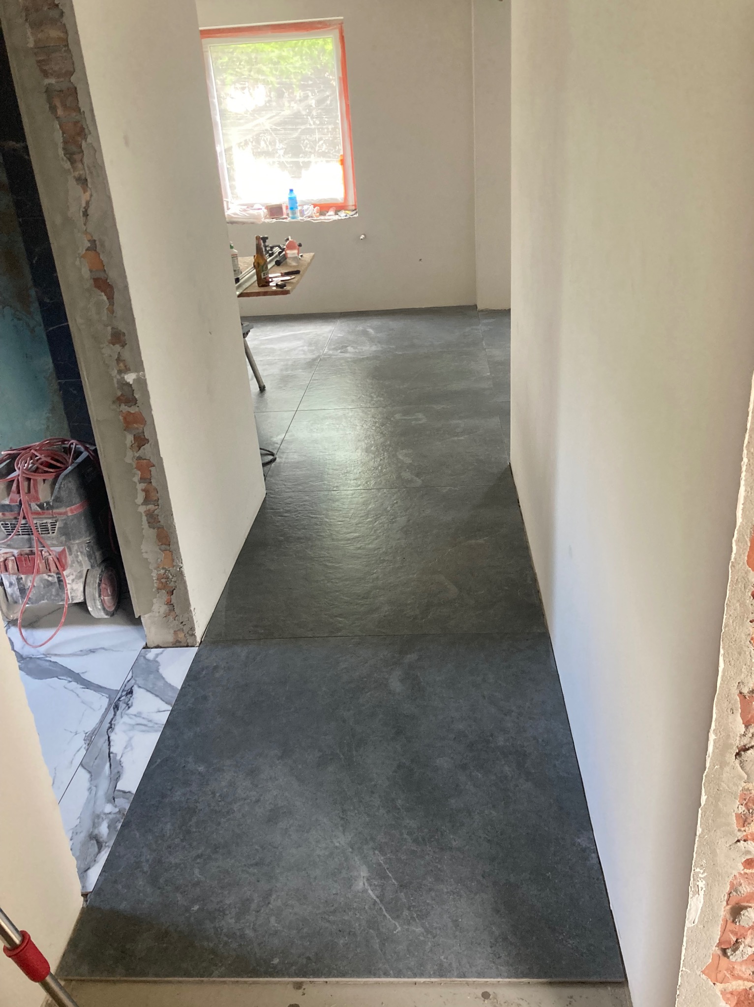 Korytarz w trakcie prac wykończeniowych: szare płytki podłogowe ułożone wzdłuż, ściany przygotowane do malowania, widoczne surowe fragmenty muru i narzędzia w tle.
