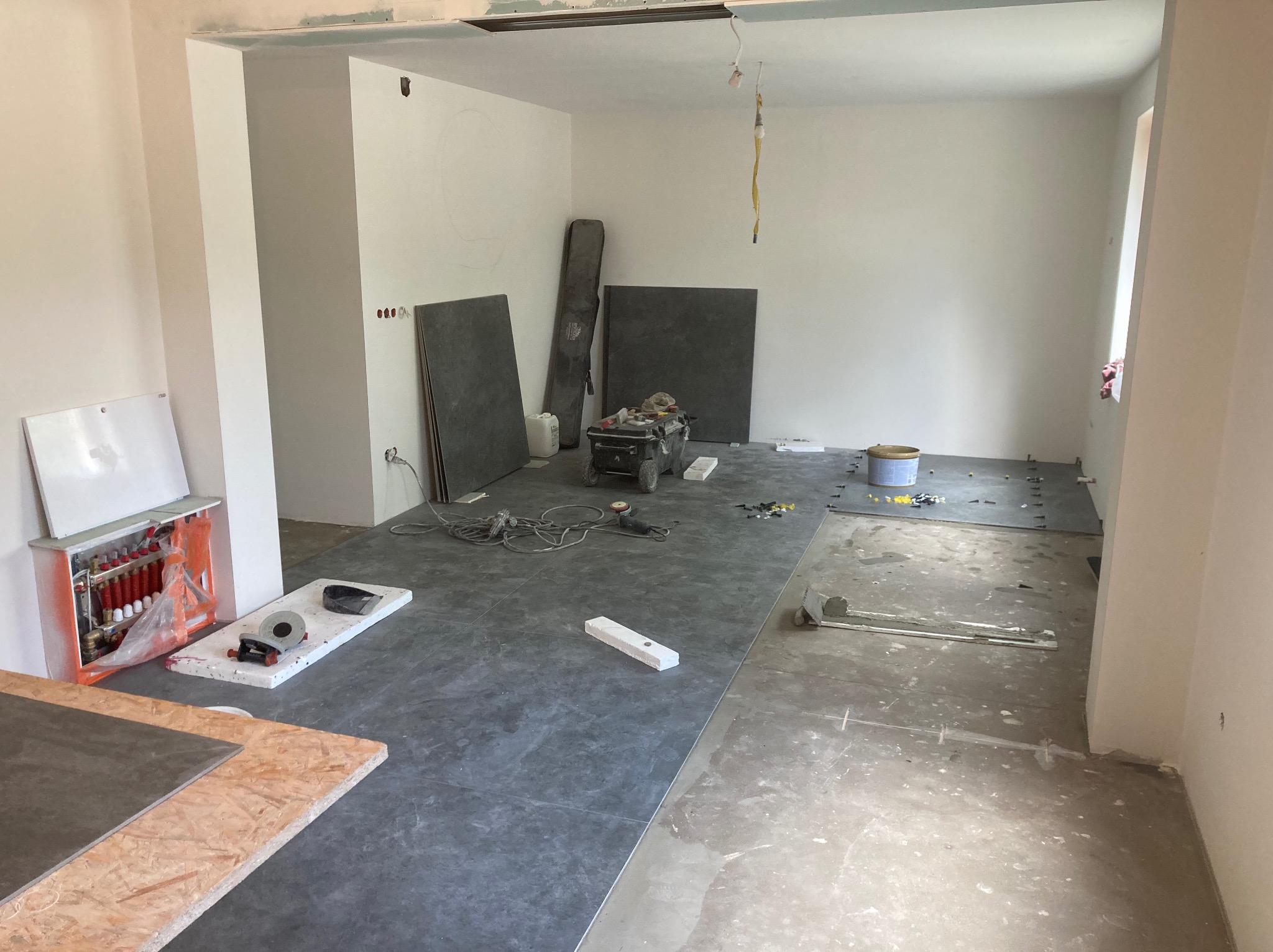 Wnętrze w trakcie układania ciemnoszarej podłogi z płytek, widoczne narzędzia, poziomica, płyty na ścianie i instalacja ogrzewania podłogowego w szafce z pomarańczowymi elementami.