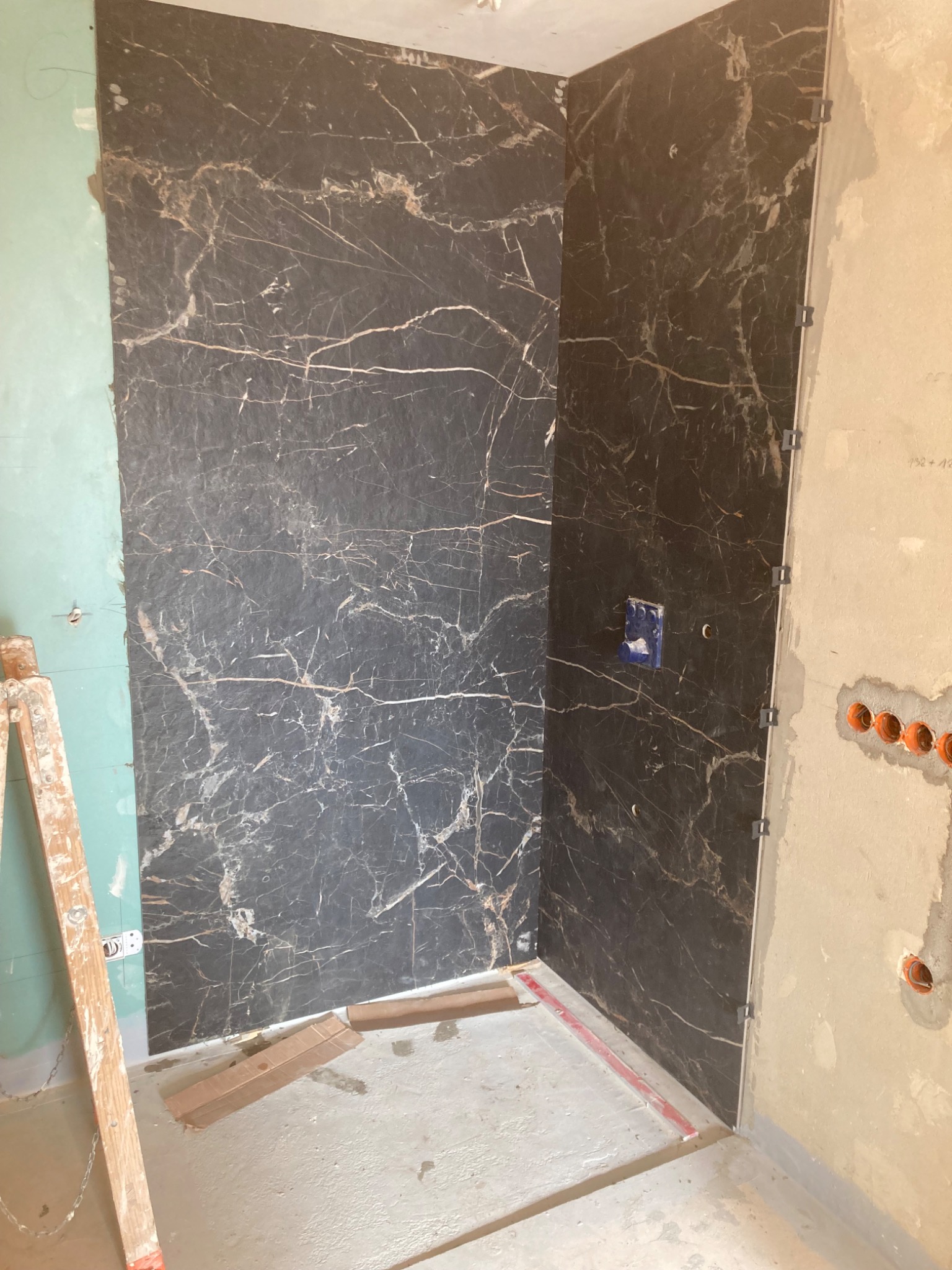 Montaż okładziny imitującej czarny marmur na ścianie w rogu łazienki, widoczne narzędzia i elementy instalacji elektrycznej, drabina oparta o ścianę z zieloną płytą.