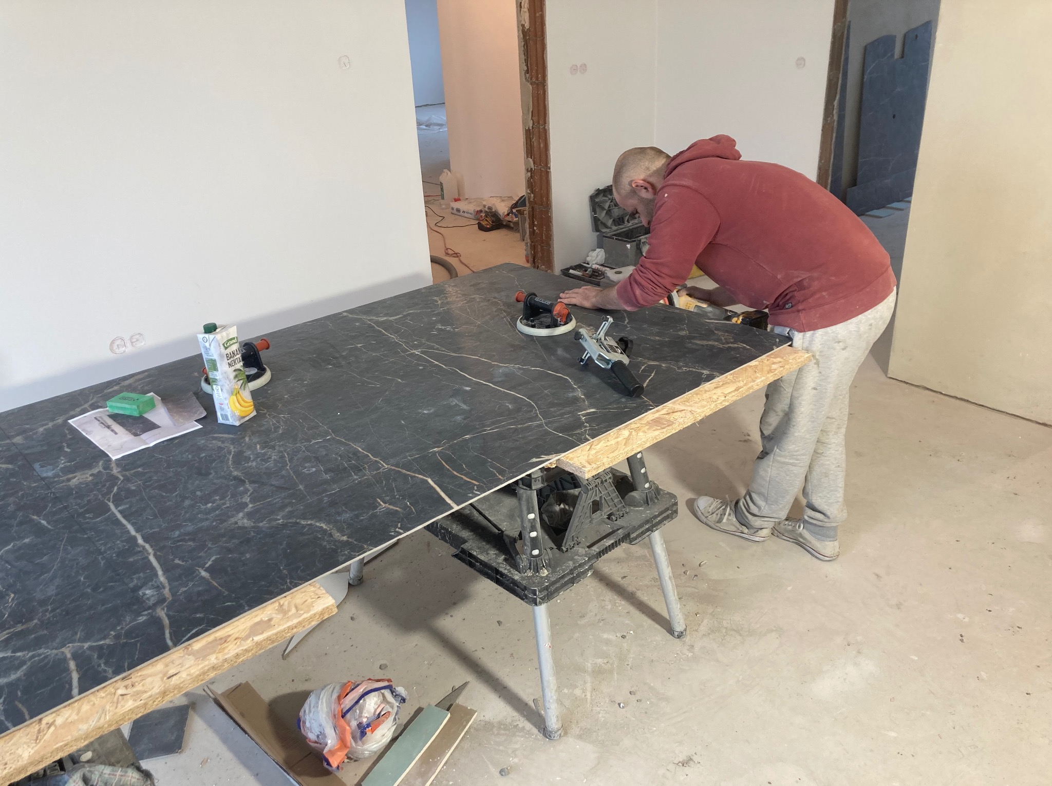 Pracownik w czerwonej bluzie obrabia płytę z czarnym wzorem marmuru na kozłach, używając szlifierki kątowej, w tle widoczne pomieszczenie w trakcie prac wykończeniowych.