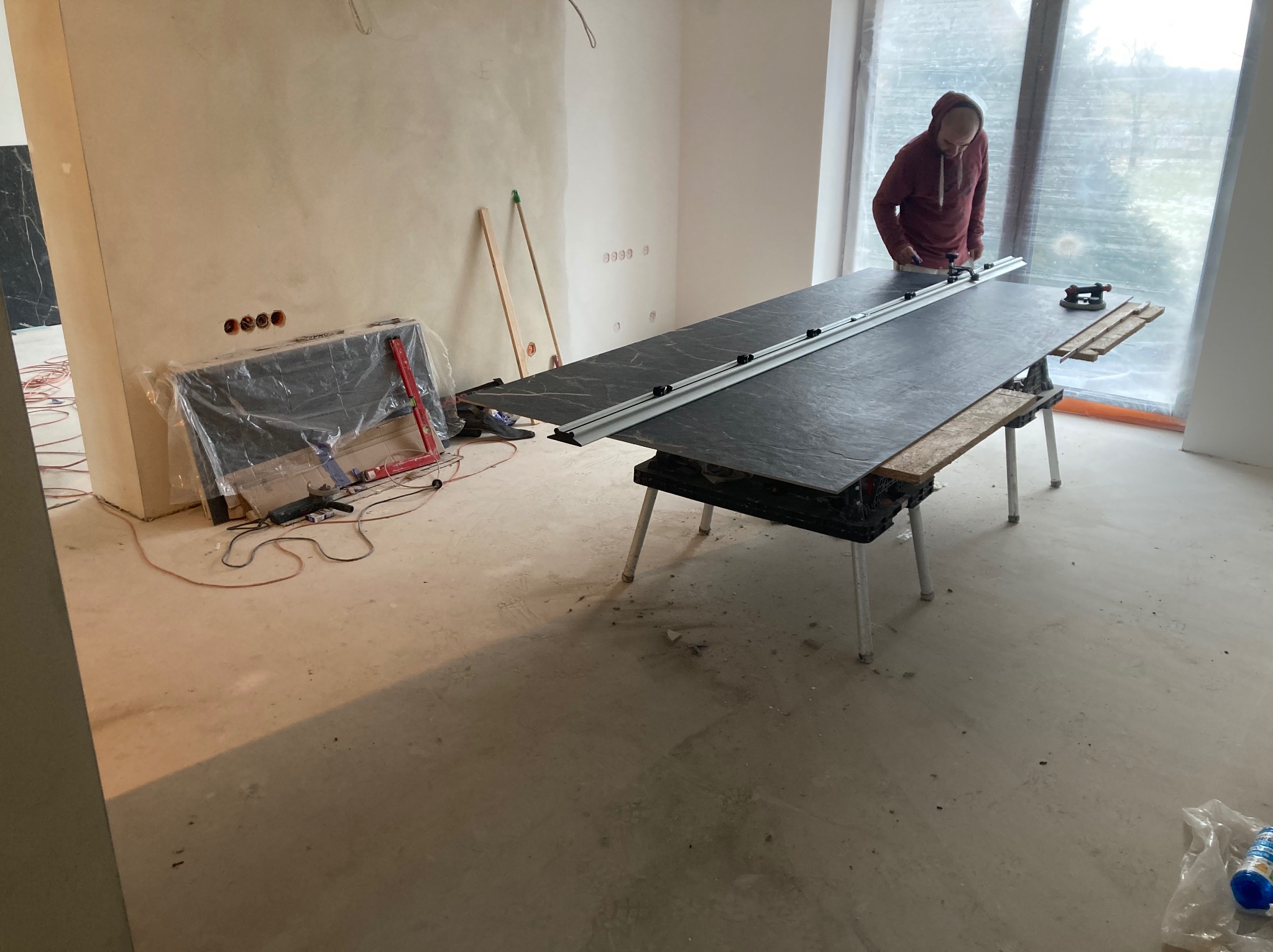 Pracownik w bordowej bluzie docina płytę z wzorem marmuru na stole roboczym w surowym wnętrzu, widoczne przygotowane ściany i okno zasłonięte folią ochronną.