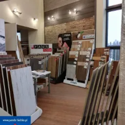 Ekspozycja paneli podłogowych w sklepie: różnorodne próbki i wzory, oświetlenie halogenowe, panele ułożone w stojakach, widoczny fragment podłogi z paneli, w tle regały z materiałami.