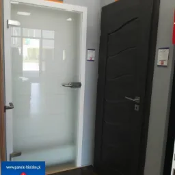 Ekspozycja dwóch modeli drzwi wewnętrznych: szklane drzwi przesuwne z białymi ramami oraz ciemne drzwi z dekoracyjnymi frezami, oba z logo DRE nad nimi.