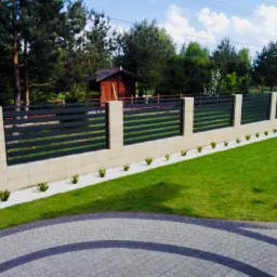 Nowoczesne ogrodzenie z poziomych, czarnych paneli w Białymstoku, osadzone na beżowych słupkach z bloczków, z białym żwirem i niskimi krzewami wzdłuż podstawy, widok z kostki brukowej.