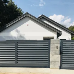 Nowoczesna, przesuwna brama wjazdowa z szarymi, poziomymi panelami i betonowymi słupkami, na tle nowego domu z szarą dachówką.