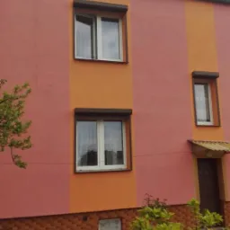 Dwukondygnacyjny dom z elewacją w odcieniach różu i pomarańczy, z oknami z brązowymi roletami, widoczne pęknięcia na ścianie, ceglana podmurówka i rabata z czerwonymi tulipanami.