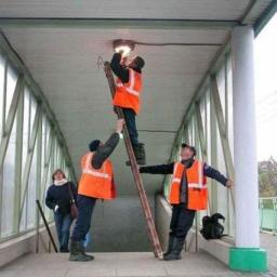 Qualius Jacek Krytwiński - Trzech mężczyzn w pomarańczowych kamizelkach wymienia żarówkę w przejściu dla pieszych, jeden stoi na drabinie, drugi ją podtrzymuje, a trzeci asekuruje balansując. Kobieta w tle wchodzi po schodach.