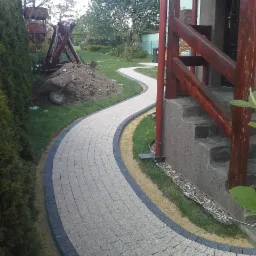 Kręta ścieżka z jasnoszarej kostki brukowej z ciemnoszarym obramowaniem, prowadząca przez zadbany ogród z elementami placu zabaw i stertą ziemi z taczką w tle