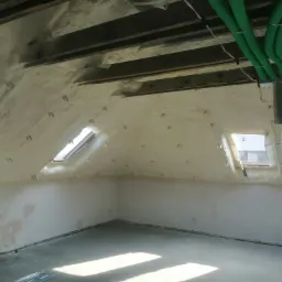 Poddasze po natrysku izolacji, widoczne okna dachowe i zielone przewody wentylacyjne pod sufitem.