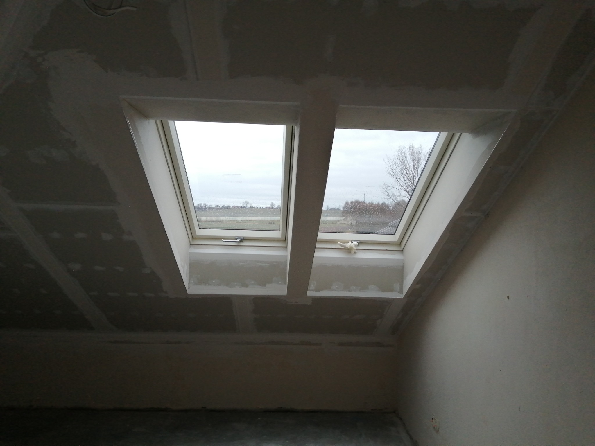 Poddasze z oknami dachowymi i płytami gipsowo-kartonowymi, gotowe do szpachlowania. Widoczny styk płyt i fragment krajobrazu za oknem.