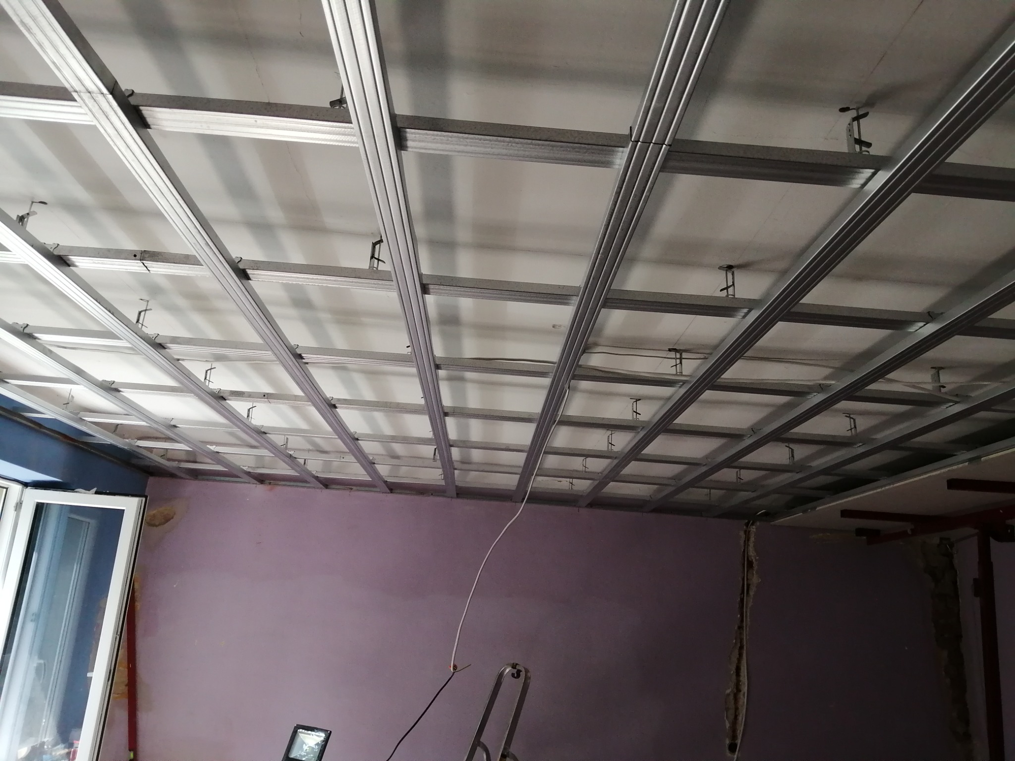 Szkielet sufitu podwieszanego z profili metalowych, widok z dołu. Częściowo zamontowane płyty. Fioletowa ściana w tle. Etap prac wykończeniowych.