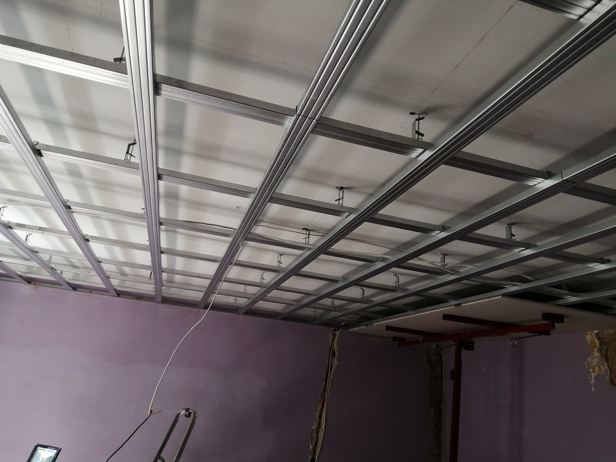 Stelaż sufitu podwieszanego z profili metalowych, widok z dołu w trakcie montażu. Fioletowe ściany w tle, fragment drabiny i podnośnika do płyt.