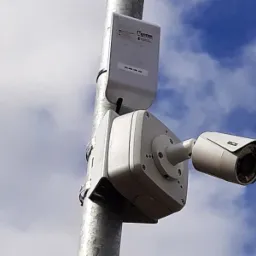 Zewnętrzna kamera monitoringu zamontowana na metalowym słupie, z widocznym modułem zasilającym i komunikacyjnym, na tle błękitnego nieba z chmurami.