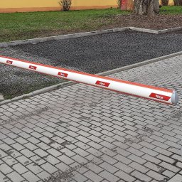 Topsystem - Szlaban parkingowy marki Nice z pomarańczową obudową i biało-czerwonym ramieniem, widoczny na tle kostki brukowej i zieleni.