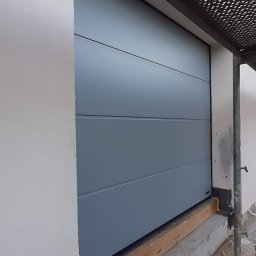 Topsystem - Szara, segmentowa brama garażowa z poziomymi przetłoczeniami, widoczna z boku, z elementami rusztowania w tle. Świeżo otynkowana ściana budynku.