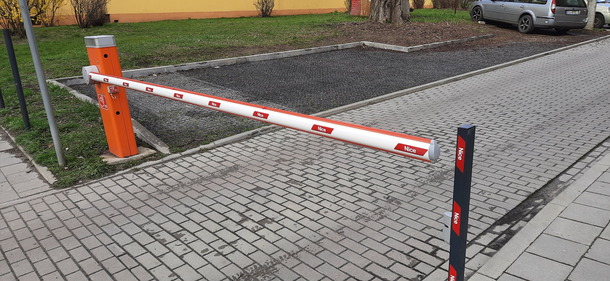Szlaban parkingowy marki Nice z pomarańczową obudową i biało-czerwonym ramieniem, widoczny na tle kostki brukowej i zieleni.