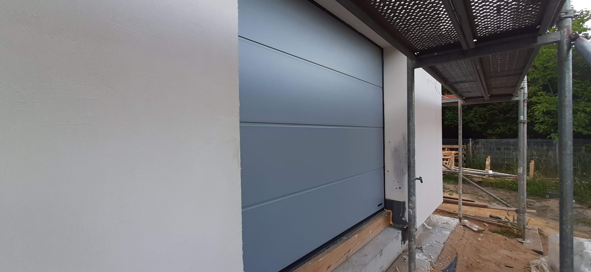 Szara, segmentowa brama garażowa z poziomymi przetłoczeniami, widoczna z boku, z elementami rusztowania w tle. Świeżo otynkowana ściana budynku.