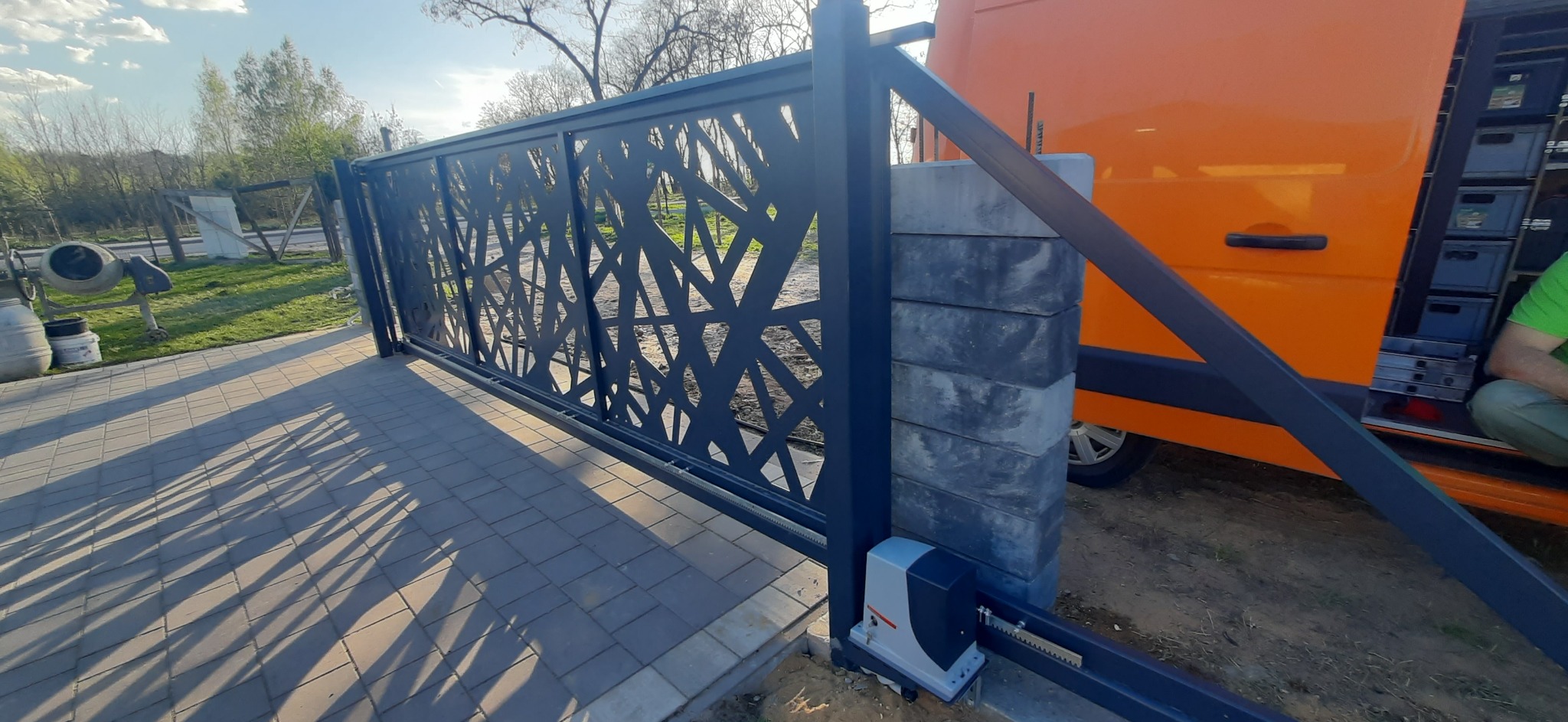 Nowoczesna brama przesuwna z geometrycznym wzorem, zamontowana na posesji. Widoczny automat do bramy i fragment pomarańczowego samochodu dostawczego.